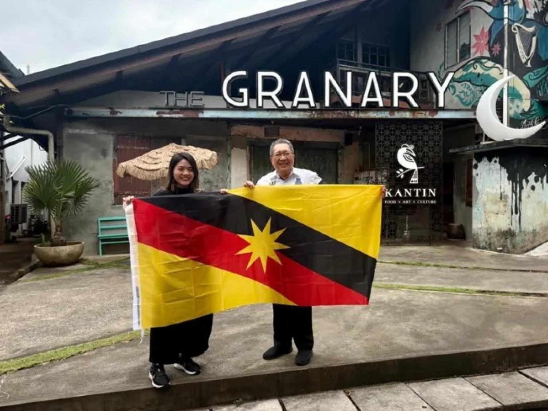 黄鸿圣（右）与KANTIN管理层Emily Yii分享喜悦时刻，并在KANTIN @ The Granary前展示砂拉越州旗。