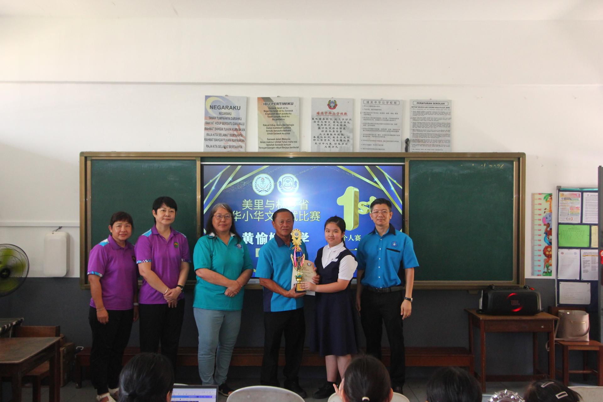 美里省与林梦省津贴华文小学董事会联合会副建设主任甲必丹严学忠（左4）颁发2026年度美里与林梦省华小华语笔试比赛个人赛第一名予美里中华公学学生黄愉婕（右2）。左起为珠巴中华公学副校长陈保諵、副校长何德香及校长郑鉥沄。右为美里省与林梦省津贴华小董联会建设主任谢家威。